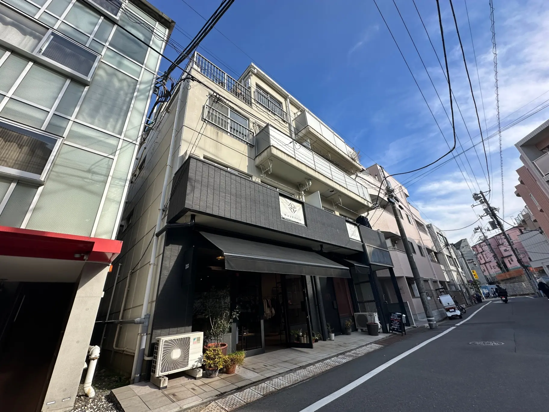 都会のビル街に位置するおしゃれな外観の店舗と周辺の建物、青空と電線が見える風景