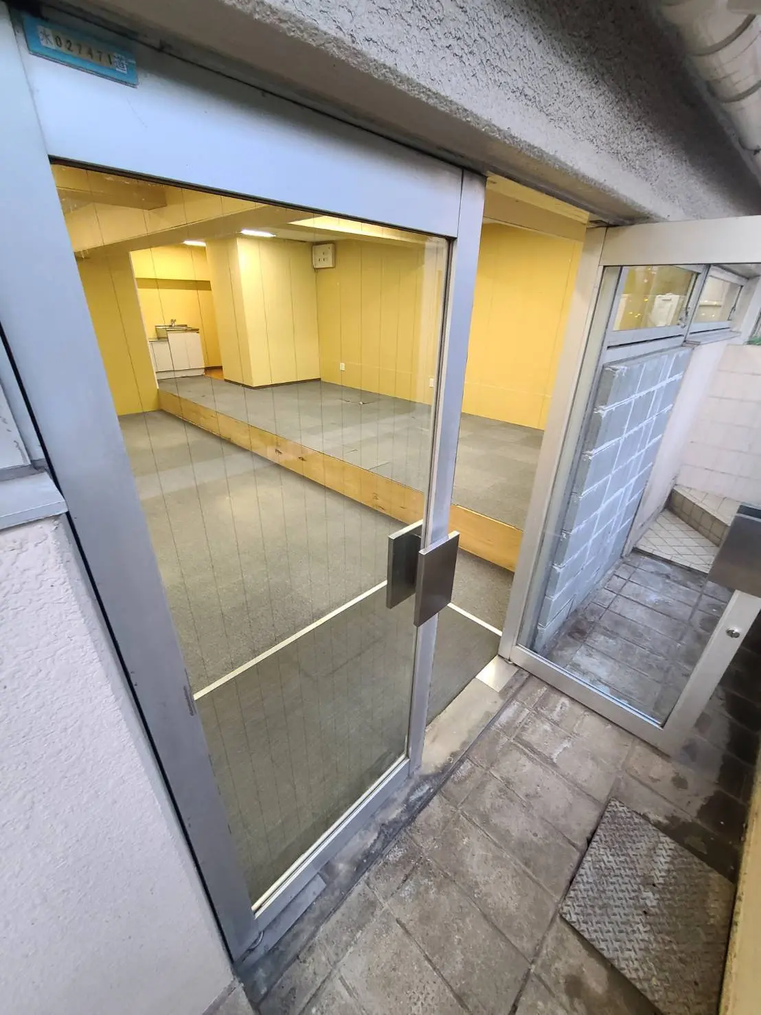 ガラス扉から見える黄色い壁の空き店舗、清潔感のある店内、石畳の歩道