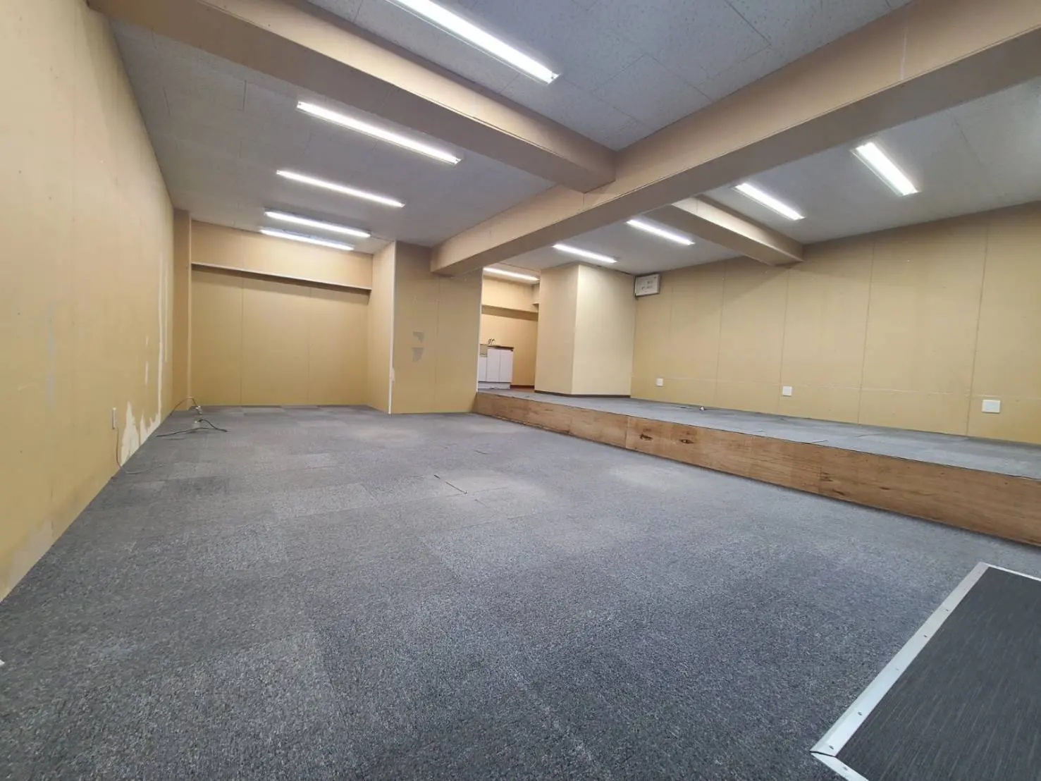 広い空間の会議室、グレーのカーペット、明るい蛍光灯、黄色い壁、木製のステージ
