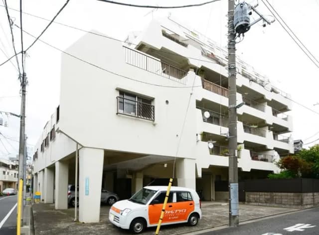- 白い外壁の4階建てマンションの外観、駐車場には小型車が停まっている様子、周囲に電柱や電線が配置されている街の風景