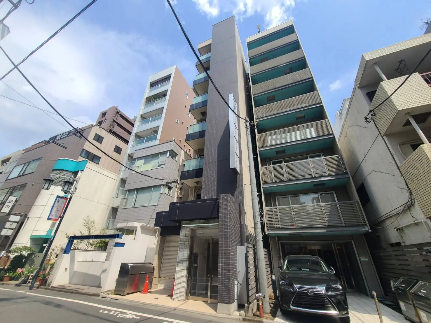 - 都会的なマンションの外観、隣接する建物との距離感、青空と共に捉えたビジュアル、1階に停まった車が目を引く