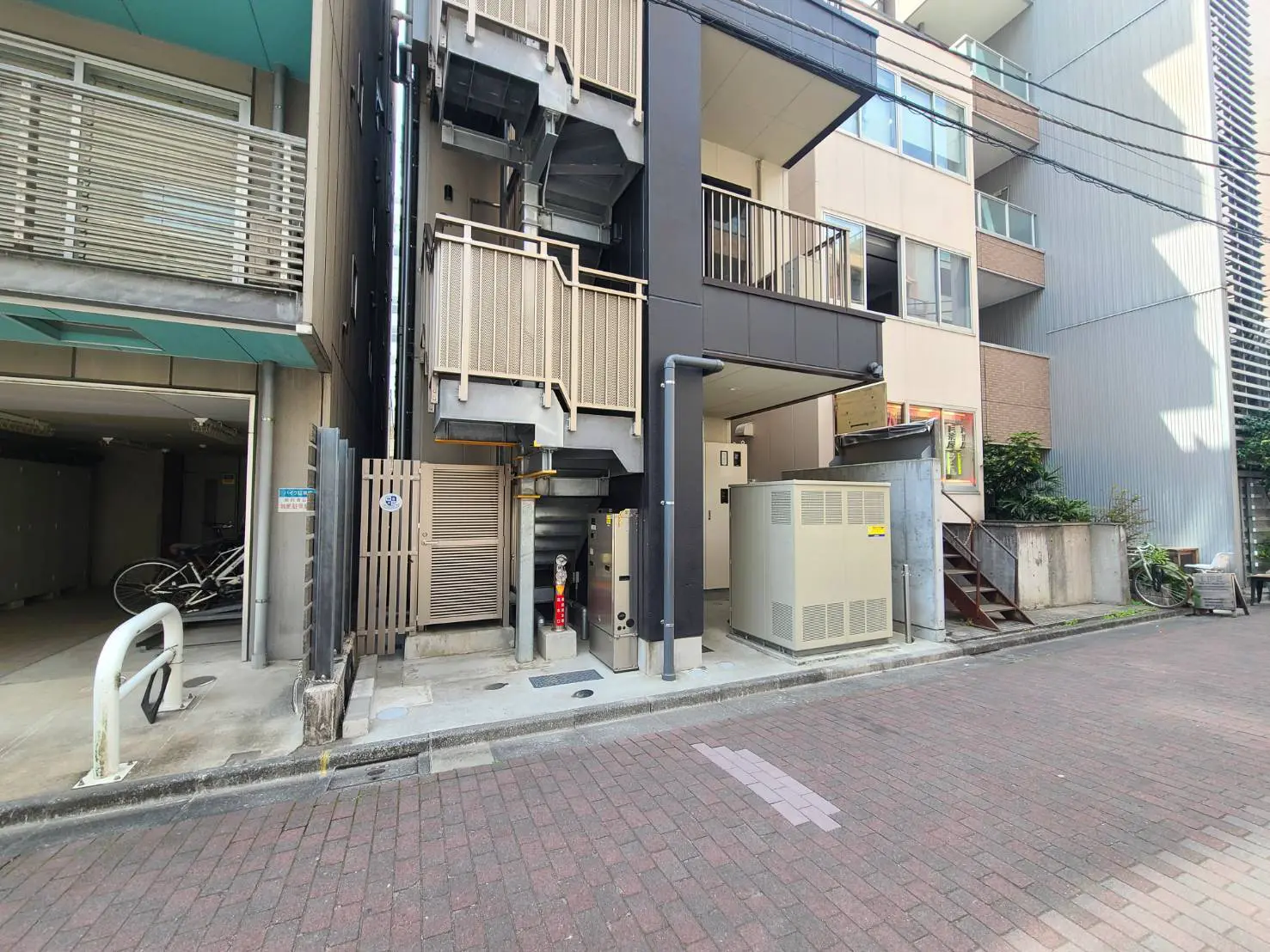 - 都市型マンションのエントランス、隣接する駐輪場と機械室、レンガ舗装の通りが見える外観