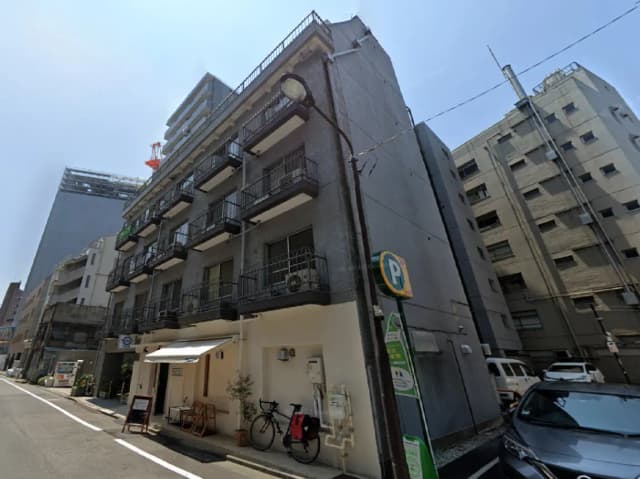 古いコンクリート造のアパートメント、バルコニーと駐輪スペースがあり、近隣に他の建物が立ち並ぶ都市風景