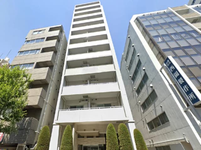 - 都市の高層ビル群の中に立つ白い外観のマンション、青空と緑の植物が映える景観