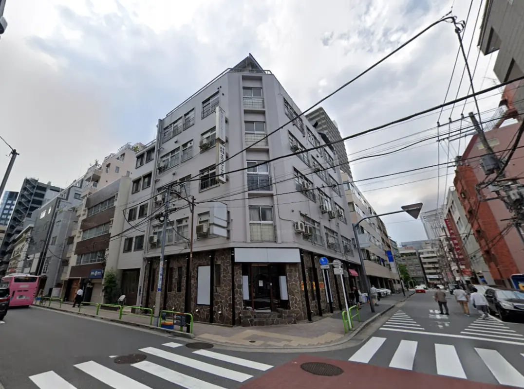 - 複数のビルが立ち並ぶ街角の風景、電線が走り、通りに人々が歩いている様子、曇り空の下の一コマ