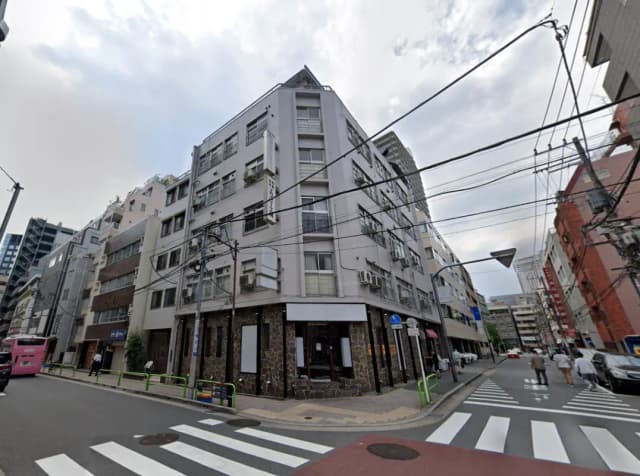 - 複数のビルが立ち並ぶ街角の風景、電線が走り、通りに人々が歩いている様子、曇り空の下の一コマ