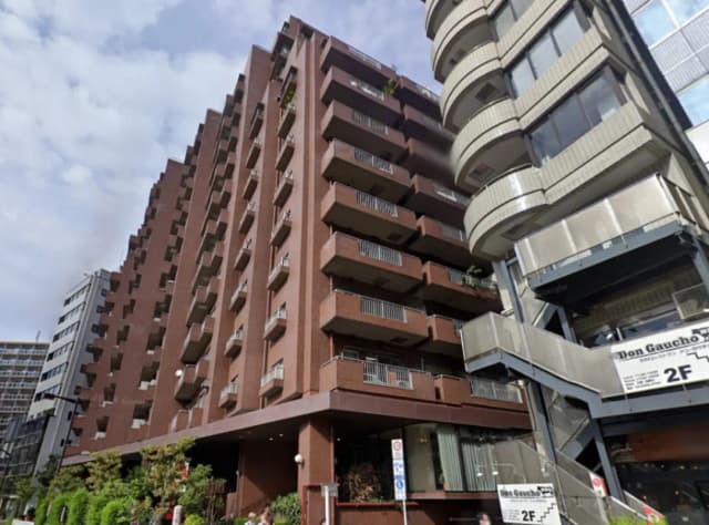 - 都市の高層ビル群の中に立つ、モダンなデザインの茶色いマンション、バルコニーと階段が特徴、周囲には植栽がある風景