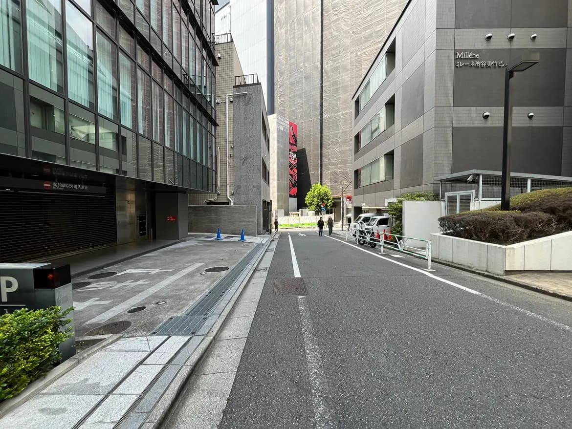 - 都市のビルに囲まれた舗装された道路、左側にはガラスのファサードの建物と駐車場のサイン、右側にはセメントの壁と緑がある街並みの風景