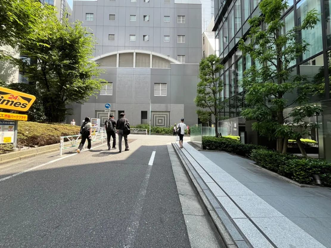 - 都市の街並みを背景に人々が歩く光景、右側には緑豊かな植栽と近代的な建物が立ち並ぶ道の風景