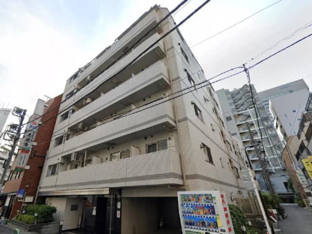 - 都市部にある4階建てのアパートメントビル、外壁は白いタイル張りで、隣接する建物が見える街の風景