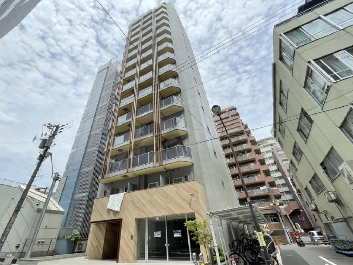 - モダンなコンクリートの外観を持つ高層マンション、周囲にはオフィスビルや住宅が立ち並ぶ街並み、青空と雲が広がる風景