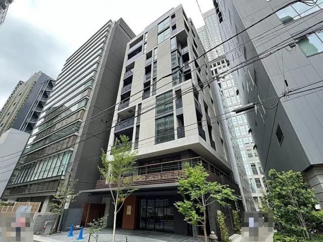 - 都市的なデザインのモダンなビル、周囲に高層ビルが並び、エントランスに緑の植栽がある様子