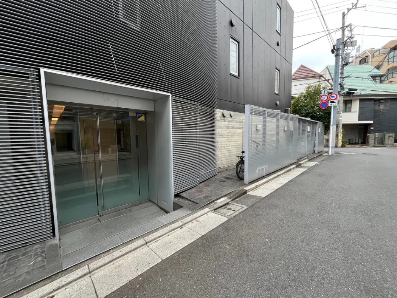 現代的な外観のビルの入り口、グレーの壁面、透明なガラス扉、周囲には住宅が見える静かな通り