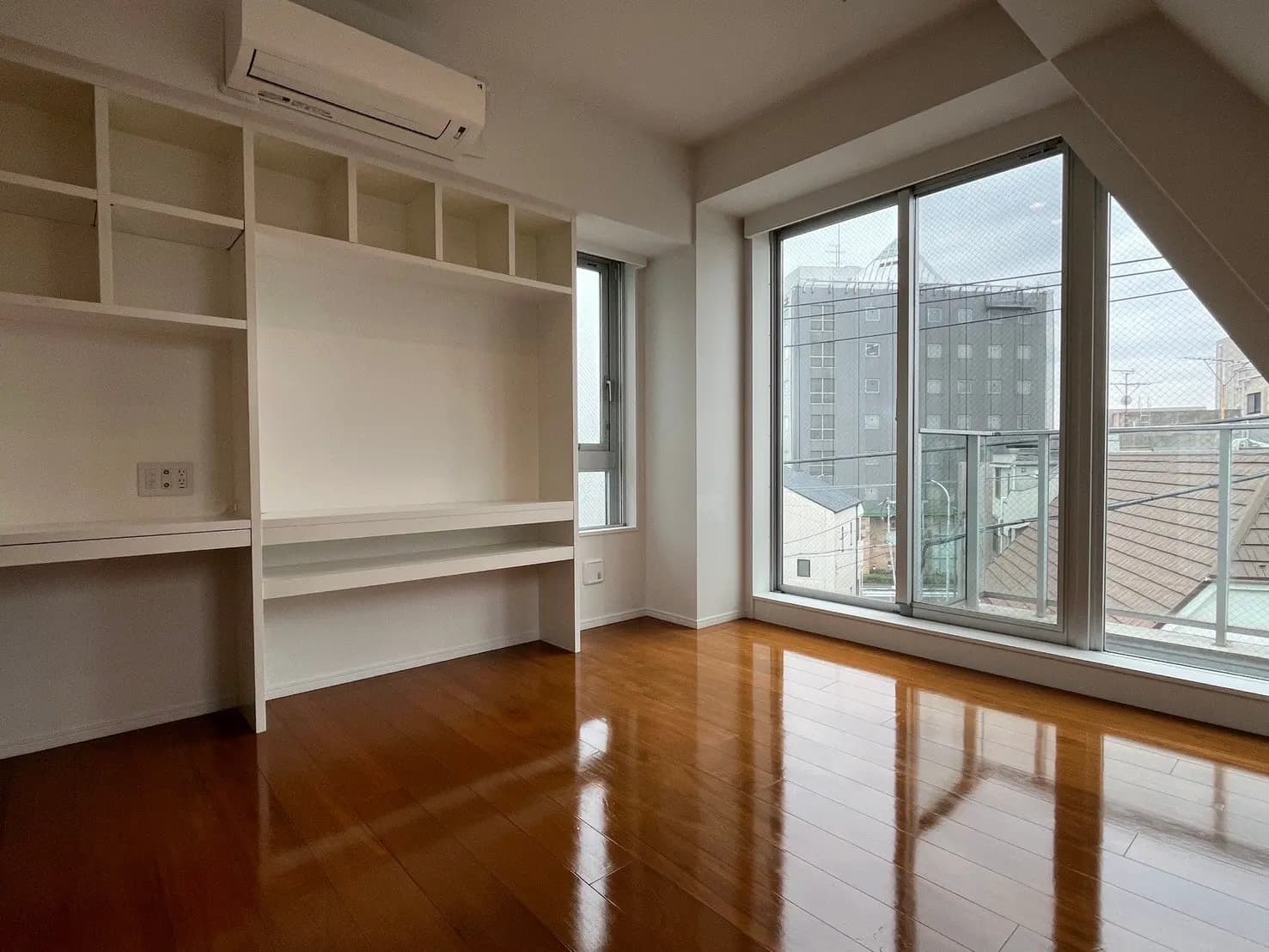 明るい自然光が差し込む広々とした部屋、木製フローリングと大きな掃き出し窓、壁には組み込み式の棚とエアコンが設置されたインテリア