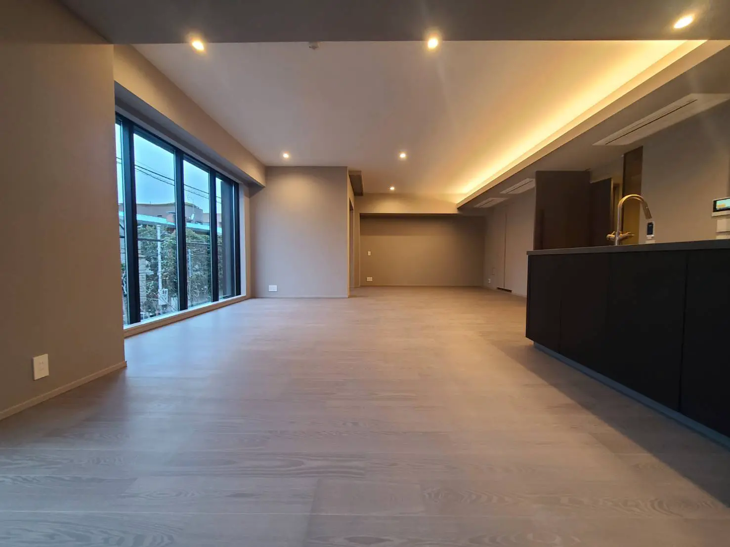 広々としたモダンなリビングルーム、天井の間接照明、大きな窓、そしてダークカラーのキッチンキャビネットが特徴のインテリア