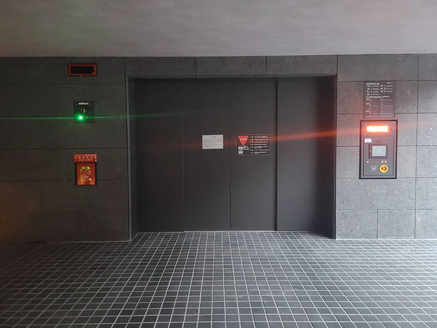 モダンな建物のエレベーターホール、灰色のタイル壁と床、緑色の動きを示すライトと非常用設備