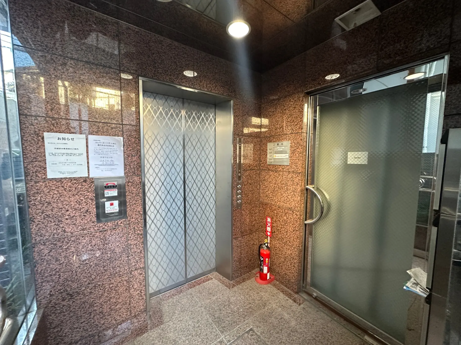 ビルのエントランス、大理石の壁と床、自動ドアと手動ドア、消火器が置かれている写真