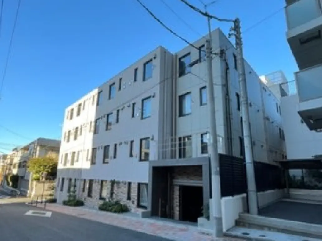 - 現代的なデザインの集合住宅外観、明るい青空の下に建つ3階建ての建物、周囲に整備された通りと植栽がある景観
