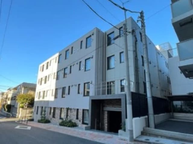 - 現代的なデザインの集合住宅外観、明るい青空の下に建つ3階建ての建物、周囲に整備された通りと植栽がある景観