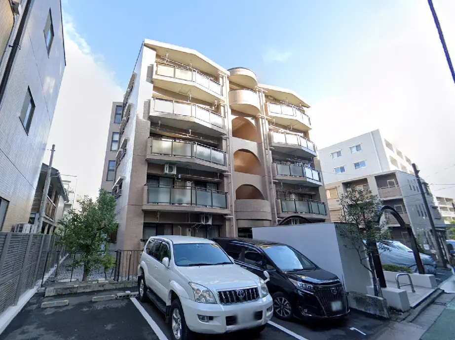 - 白い車と黒い車が停車する駐車場の前に立つ、モダンなデザインのマンションの外観、複数階建てでバルコニー付きの特徴的な建物