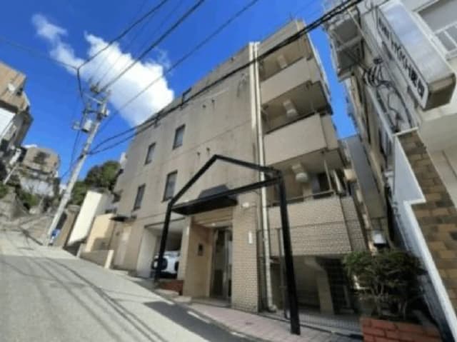 - 傾斜のある道路に面したアパートの外観、青空と白い雲が広がる背景、入り口にはシンプルな屋根状のサインがある。