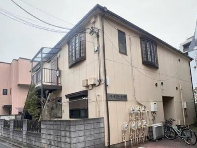 - ホワイトカラーの外壁を持つ二階建て住宅、バルコニーや装飾的な窓が特徴的で周囲に自転車や設備が見える景観