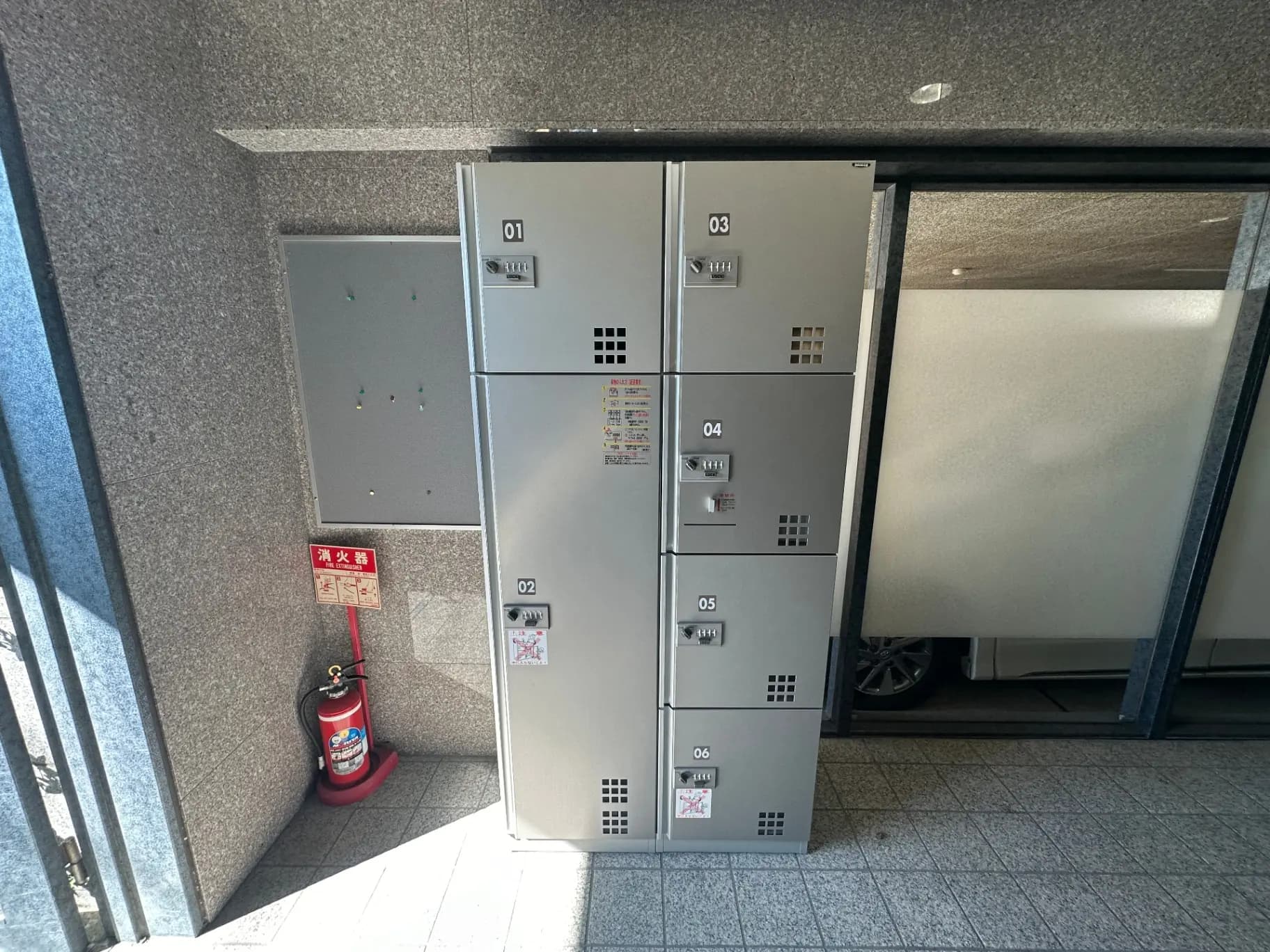 アパートメントのエントランスホールに設置された銀色の宅配ボックス、右側には消火器が置かれている光景