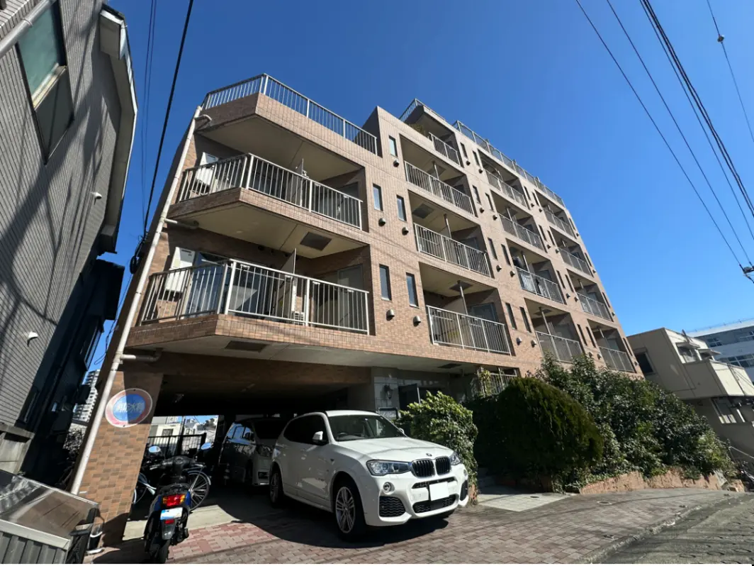 新築マンションの外観、明るい日差しの中でベージュ色の壁とベランダ、地下駐車場、道路には自転車と白い車