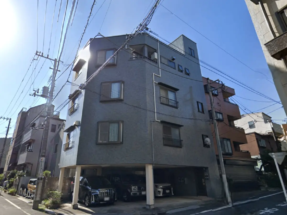 - 青空の下、電線が張られた街並みの一角に佇むモダンなマンション、1階には車が停められている様子