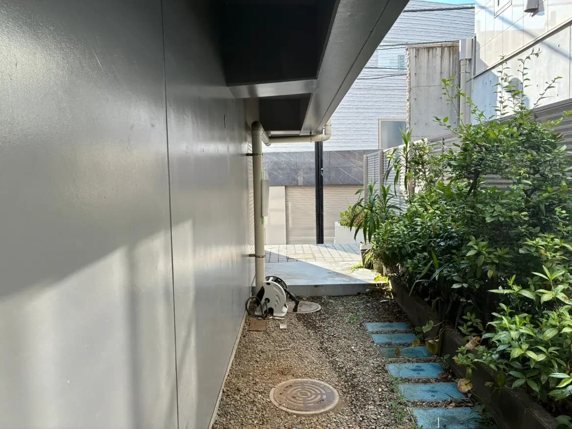 - モダンな住宅の側面、グレーの壁の隣に石の敷かれた通路と植物が生い茂る風景