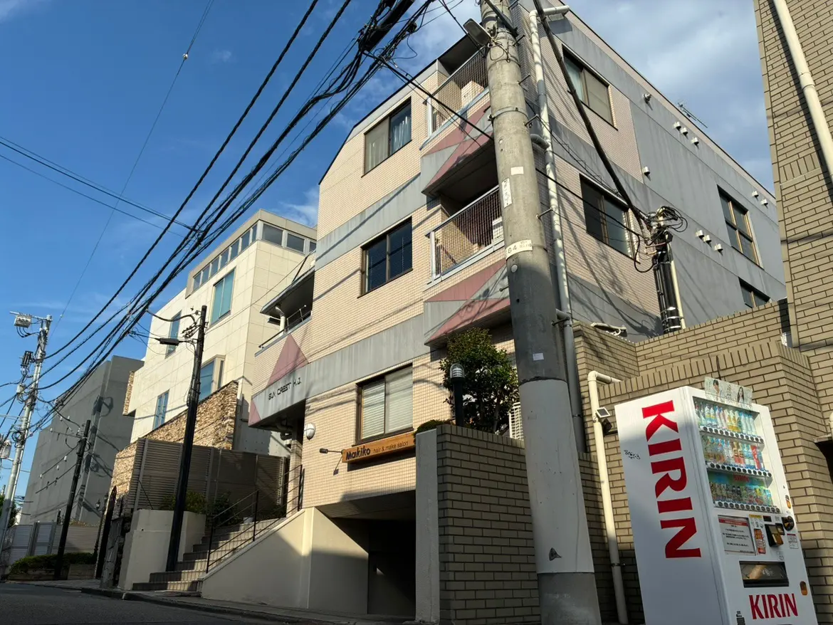 - 近代的な外観のアパートメントビル、周囲には電柱や配線、目の前には自動販売機が設置された通りの風景