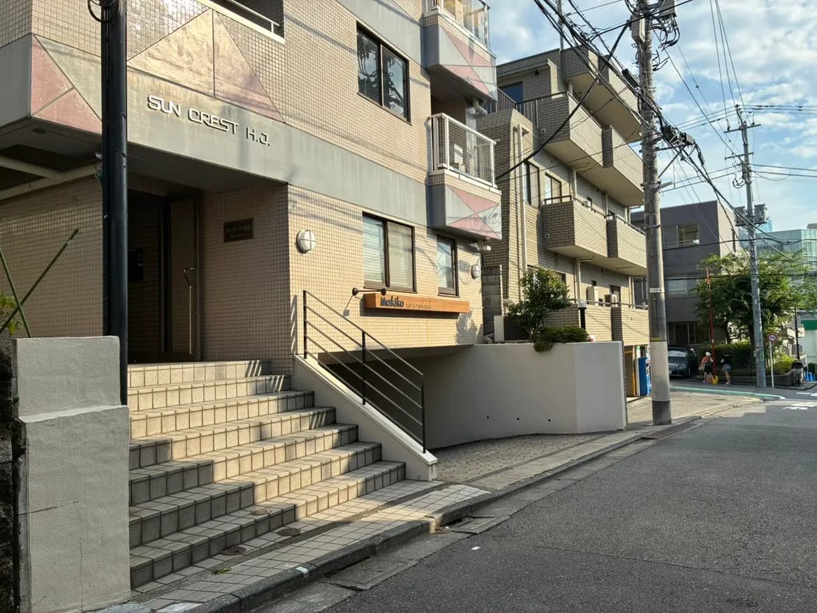 - 住宅地にある「SUN CREST H.J.」のエントランス、階段の上に位置するシンプルなデザインの建物と周囲の街並み