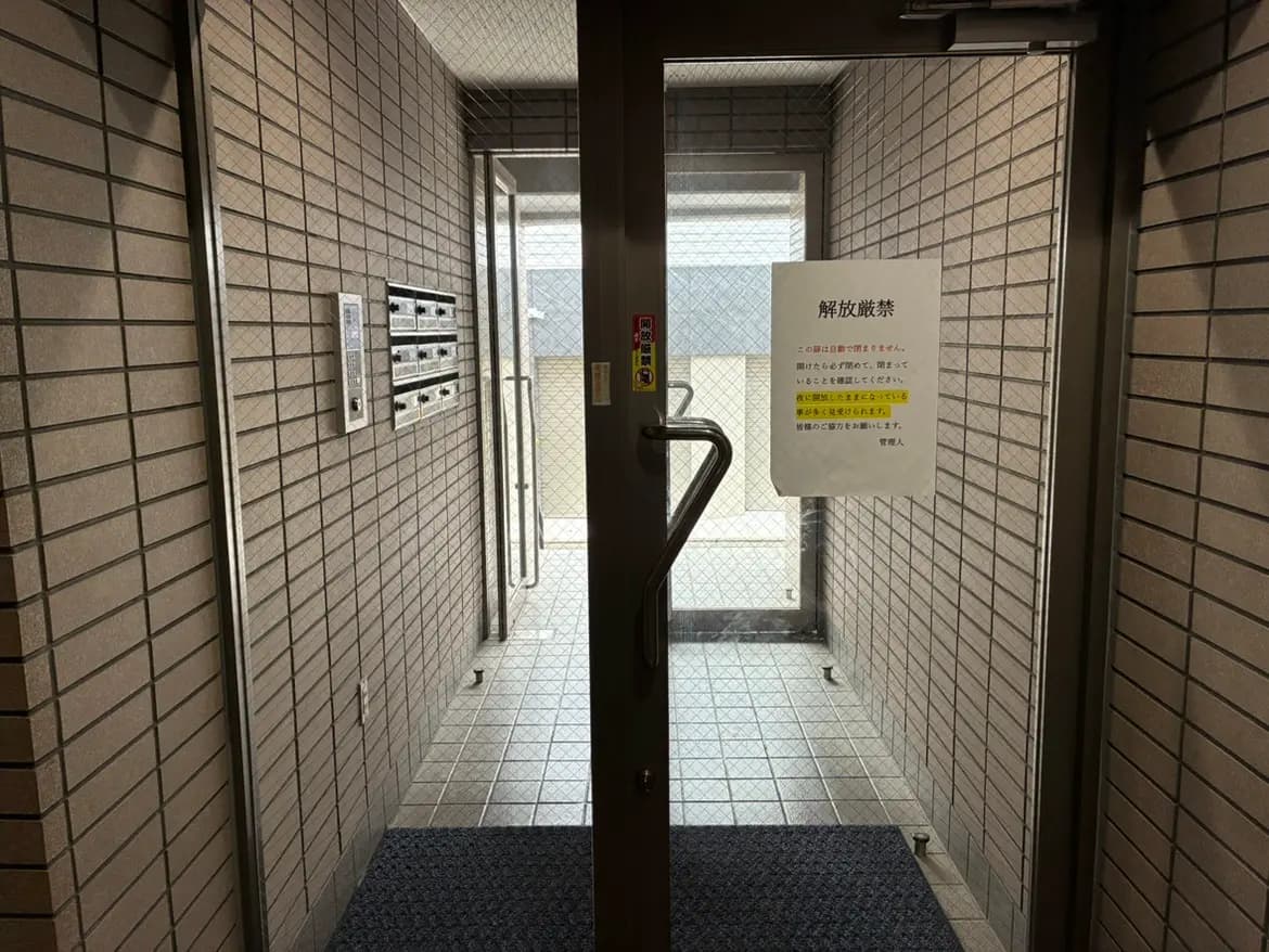 - 入口ドアとロビーの様子、壁に郵便受け、解放厳禁の注意書きが貼られた異なる材質の床タイルのインテリア