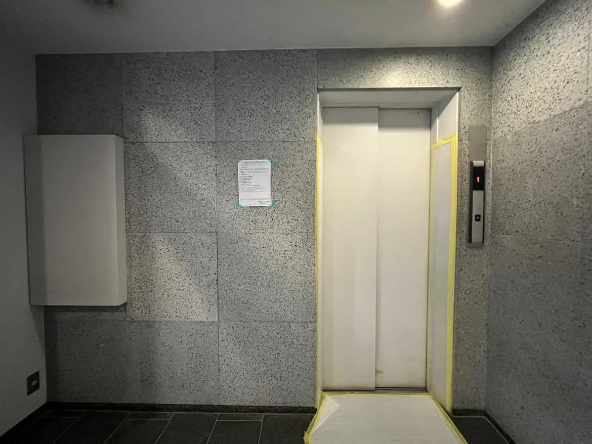 モダンな建物のエレベーター入口、グレーの壁面、黄色い注意テープ、床案内表示