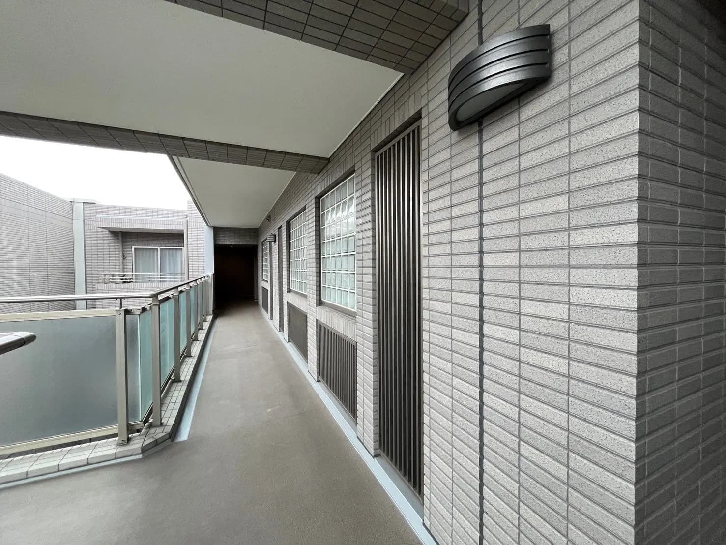 マンションの共有バルコニー、グレーのタイル壁、手すりと照明が備えられた通路