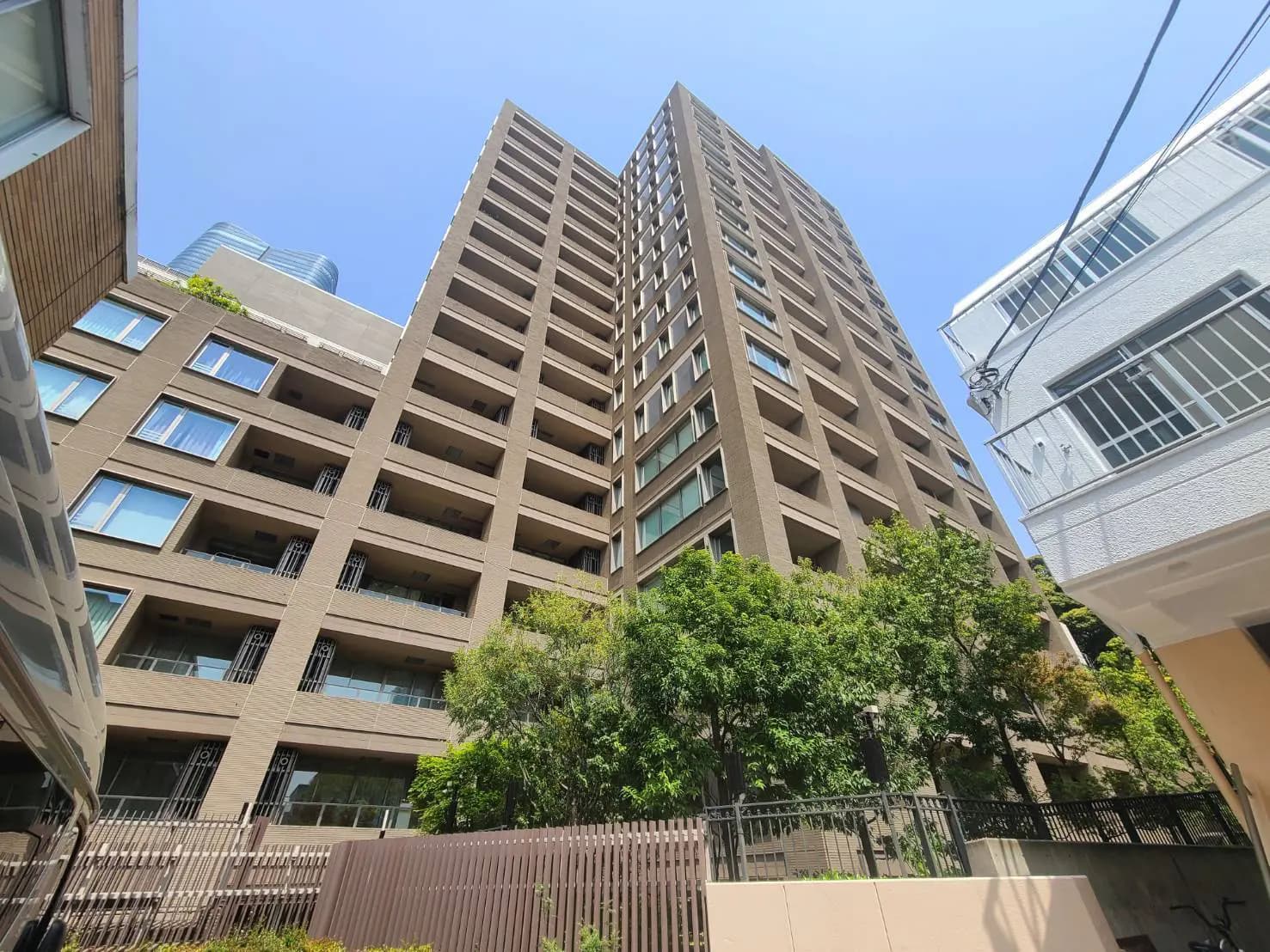 - 都市の高層マンション、青空の下、高い木々とガーデンビューテラスが隣接するエクステリア