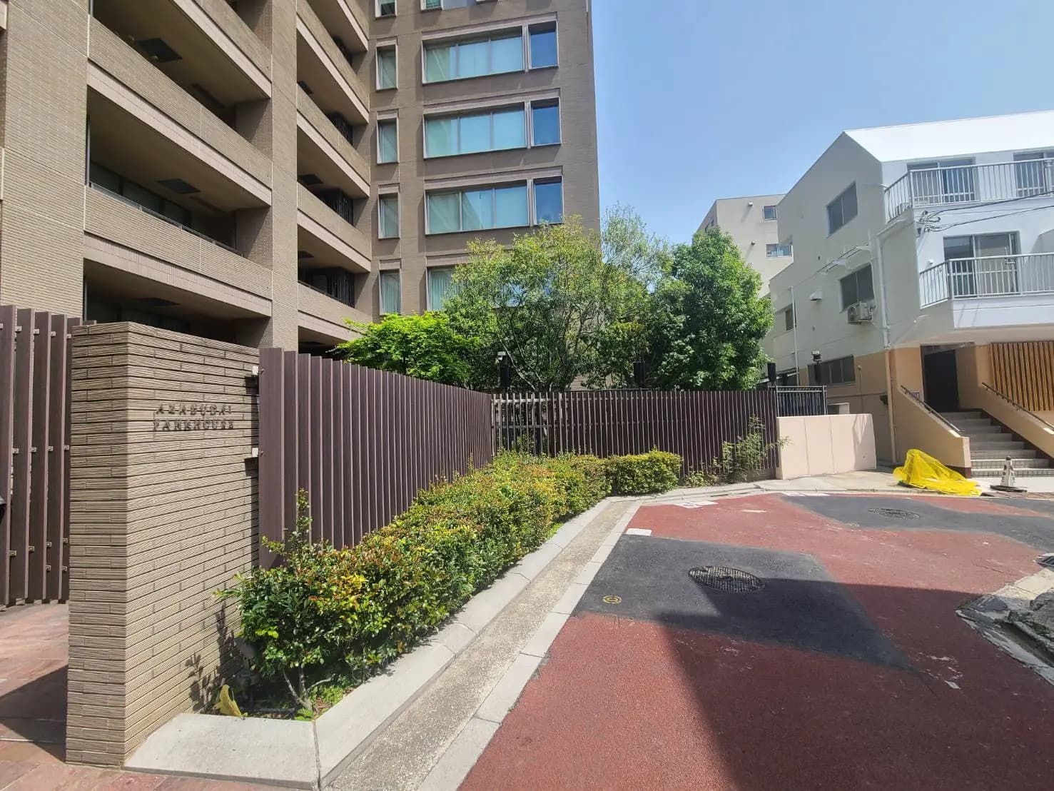 - 都市型マンションの入口風景、緑豊かな植栽と洗練されたフェンスに囲まれたエリア、近代的な建物の外観