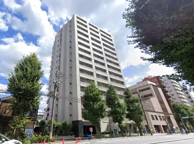 - 高層マンションの外観、周囲に並ぶ太い木々と晴れた青空が広がる風景