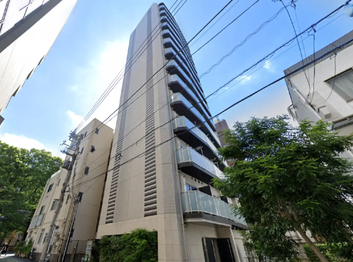 - 高層マンションの外観、近隣の建物や電柱が映り込む姿、青空と緑の木々が背景の都市風景