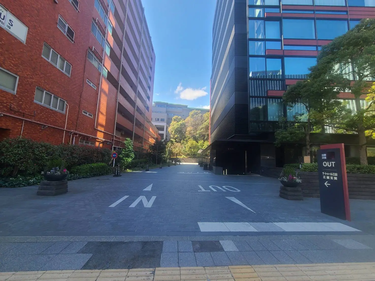オフィスビルのエントランスで「IN」と「OUT」の標示がある通路、晴天の下で赤レンガとガラス張りの建物が並ぶ風景