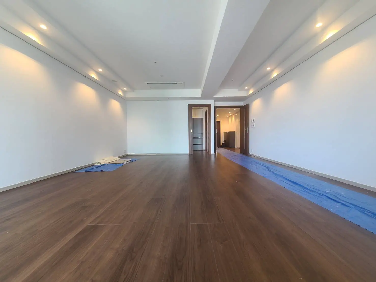 明るい照明が設置された広々とした空の部屋、木目調の床、白い壁、奥にドアが見えるインテリア