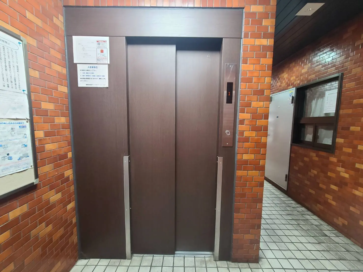 茶色い木製のドアが特徴のエレベーター前、レンガ壁と情報掲示板がある公共施設の内装