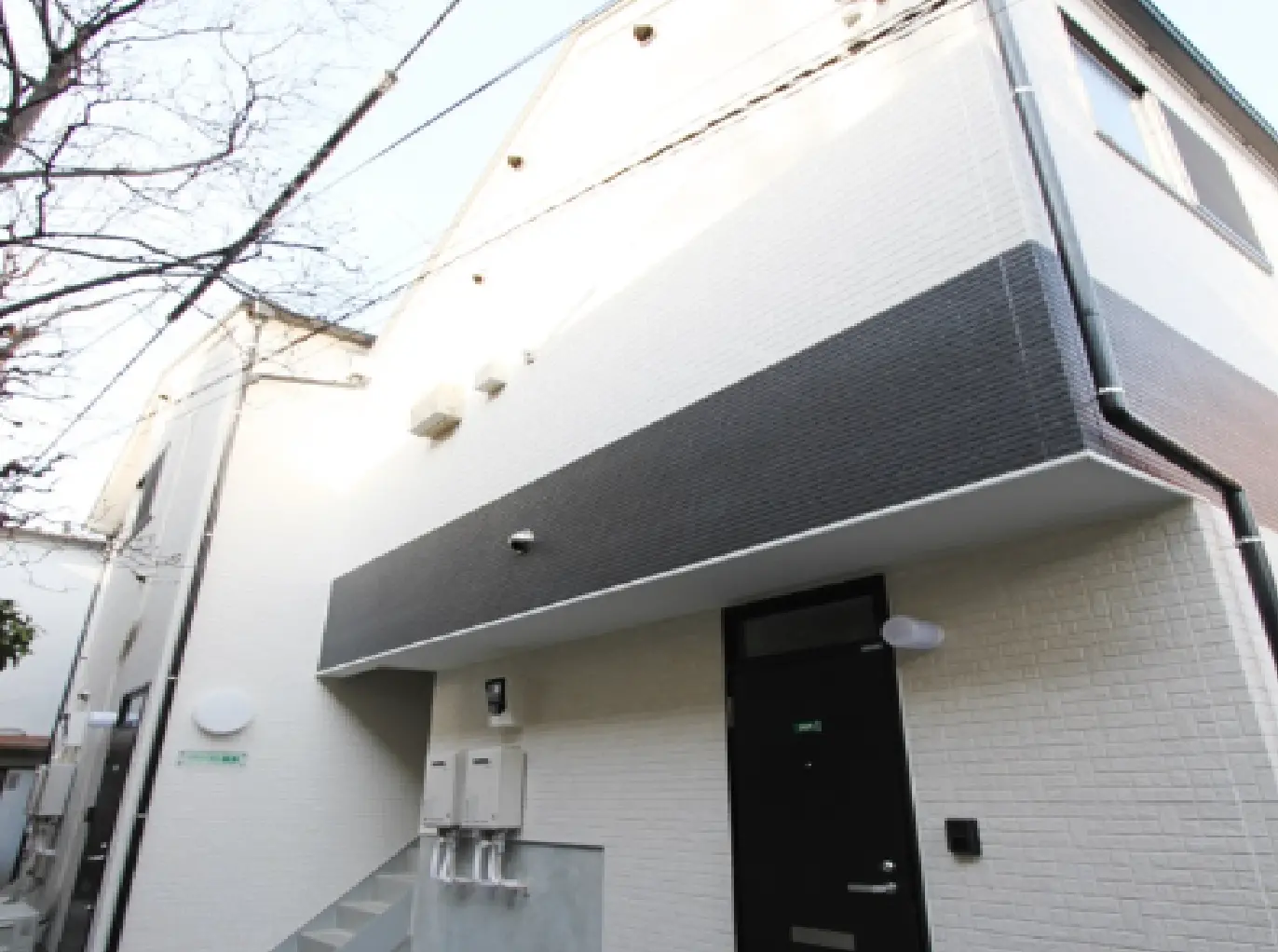- 白と黒のシンプルな外壁デザインの建物、階段とエントランス、周囲に細い電線が見える外観