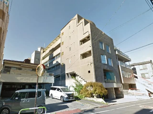 - 都市型マンションの外観、コンクリートのデザインと大きな窓が特徴的な建物、周囲に駐車スペースと緑が見える風景