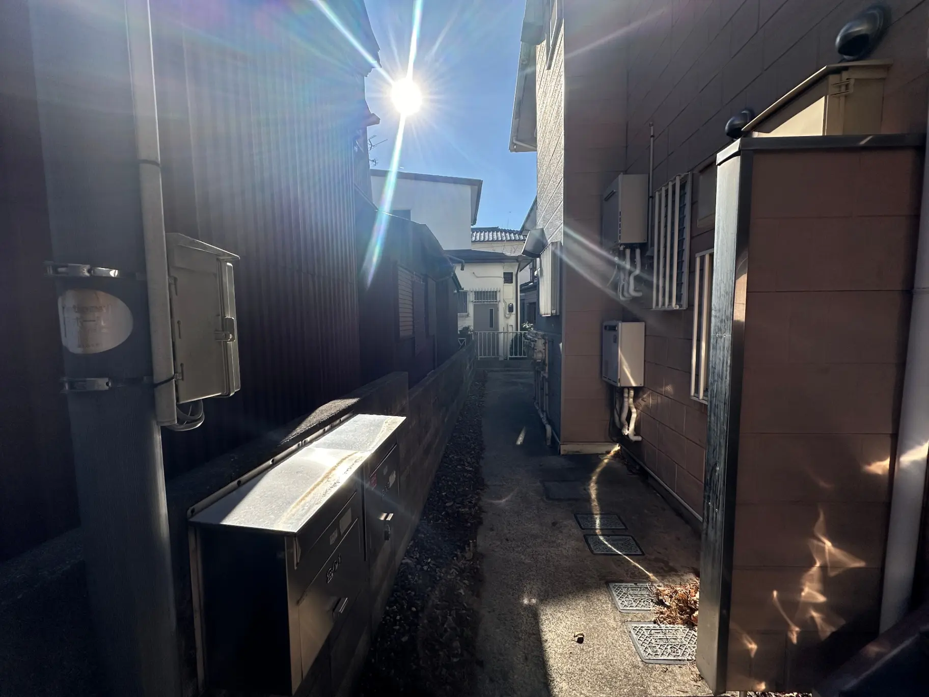 日本の狭い路地、朝日が差し込む風景、郵便受けや家の壁が写されている
