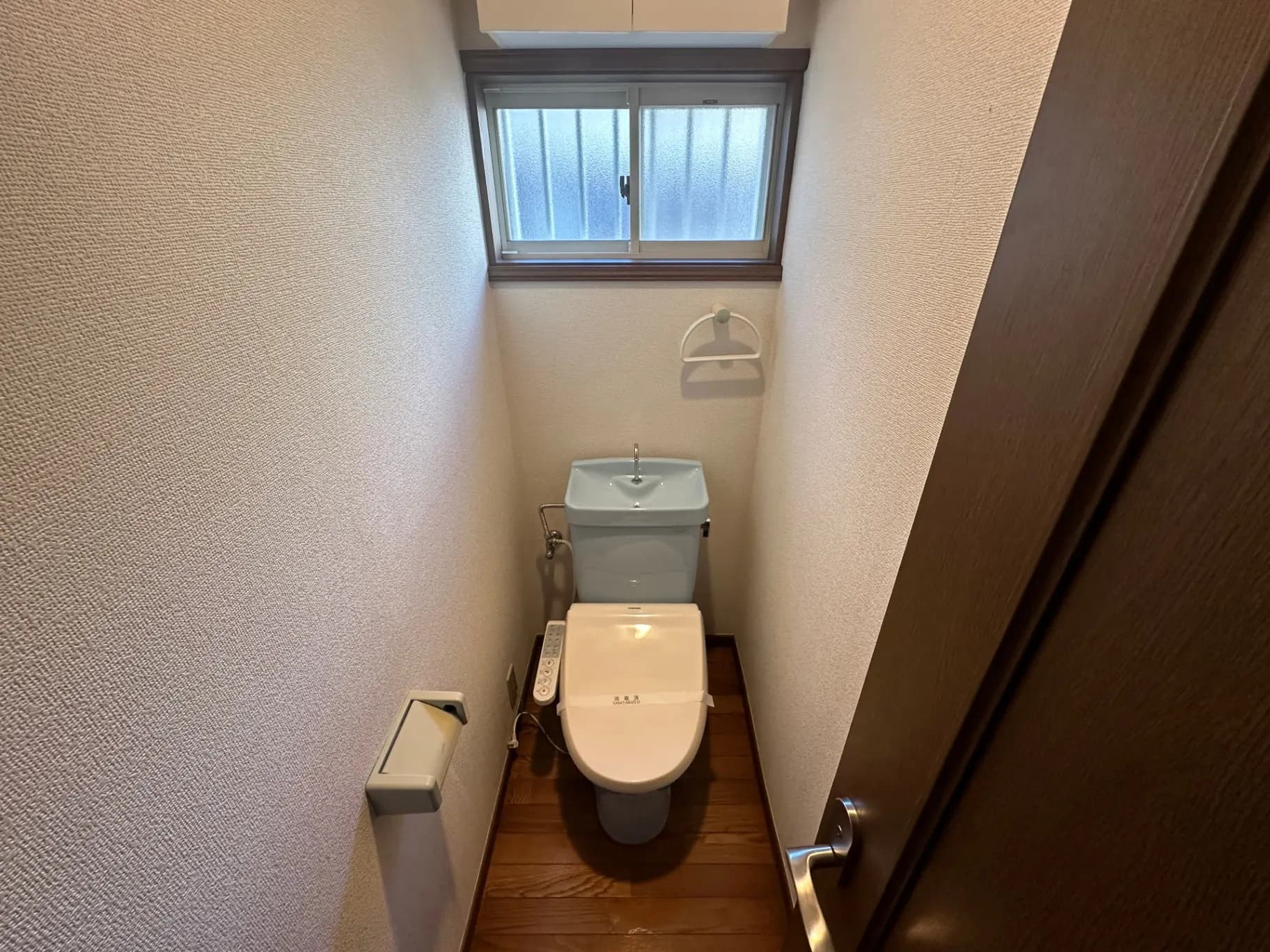 狭いトイレの内装、上部に小窓があり木製ドアとトイレットペーパーホルダーがあるシンプルなデザイン
