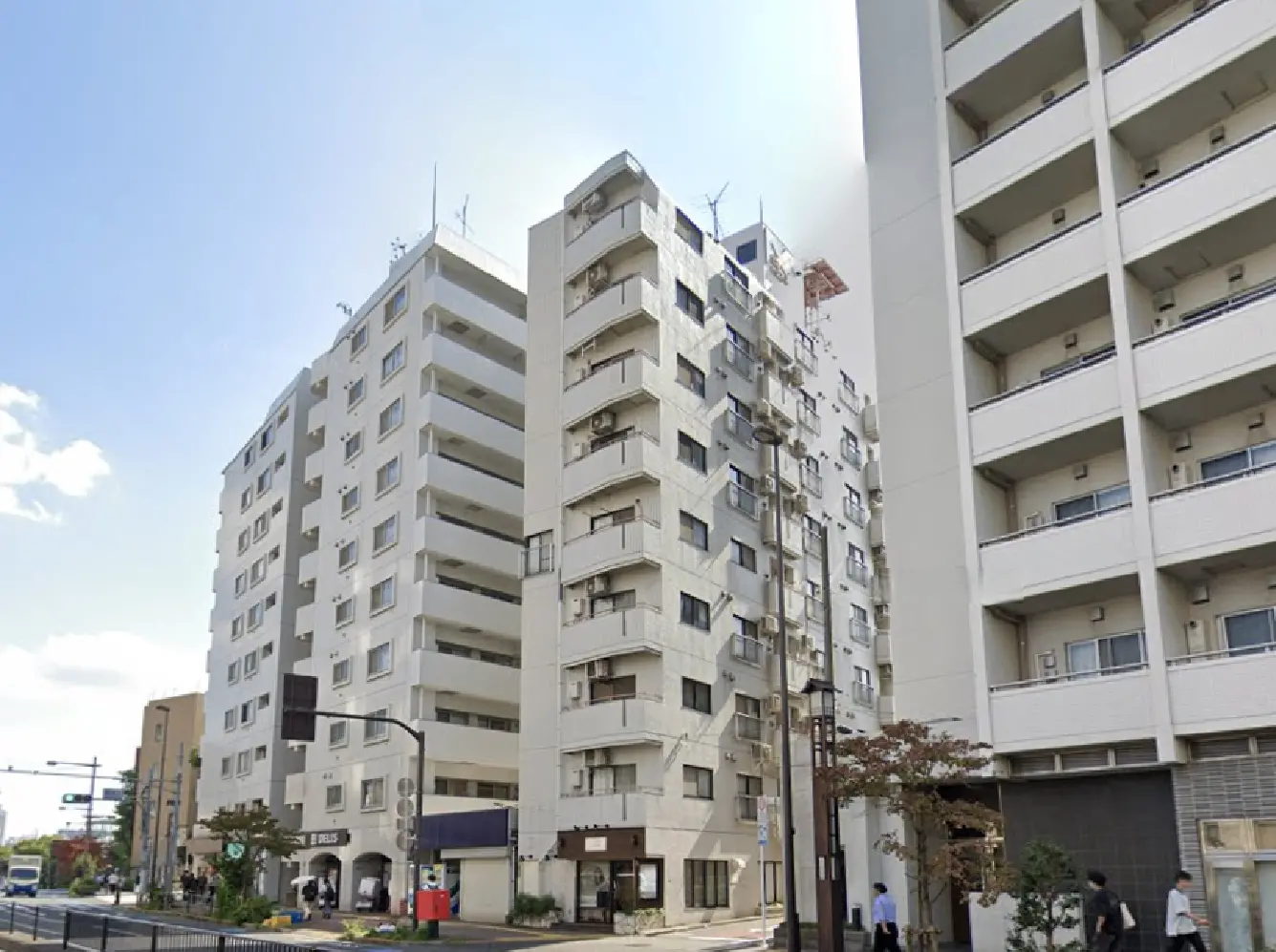 - 都市部に立つ高層マンション群、隣接する商業施設や公道が見える風景、青空と雲が広がる景観