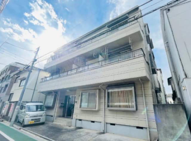- 日光が差し込む3階建てのアパート外観、周囲に住宅や街灯が見える、明るい青空と白い雲のコントラスト