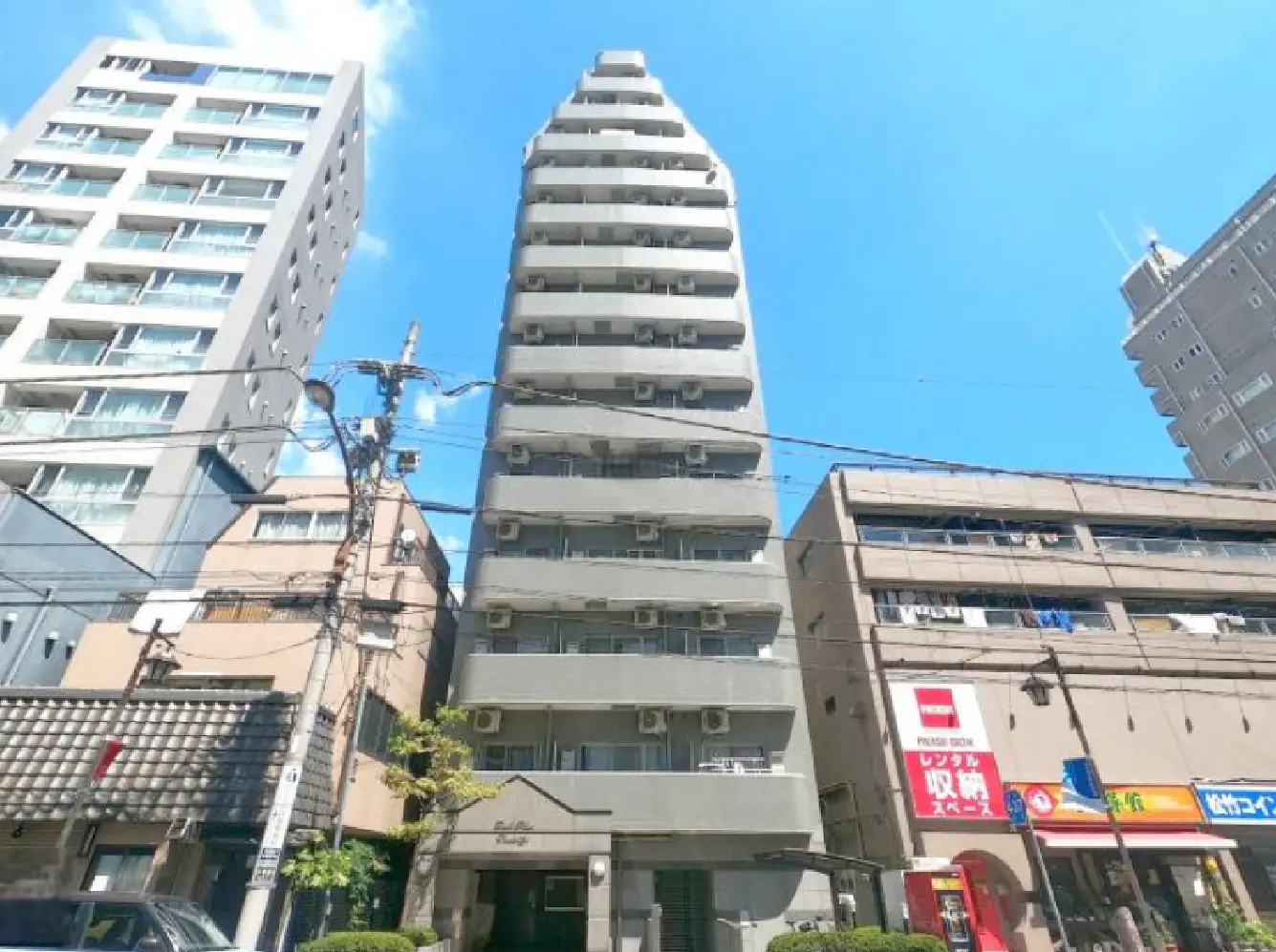 - 都市部に立つモダンなマンションビル、晴れた青空と電線が映える背景、周囲に他の建物が立ち並ぶ景観