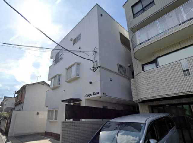 - 白い外観のアパート「Corpo Kato」、隣接する建物と敷地内の車が見える通りの風景